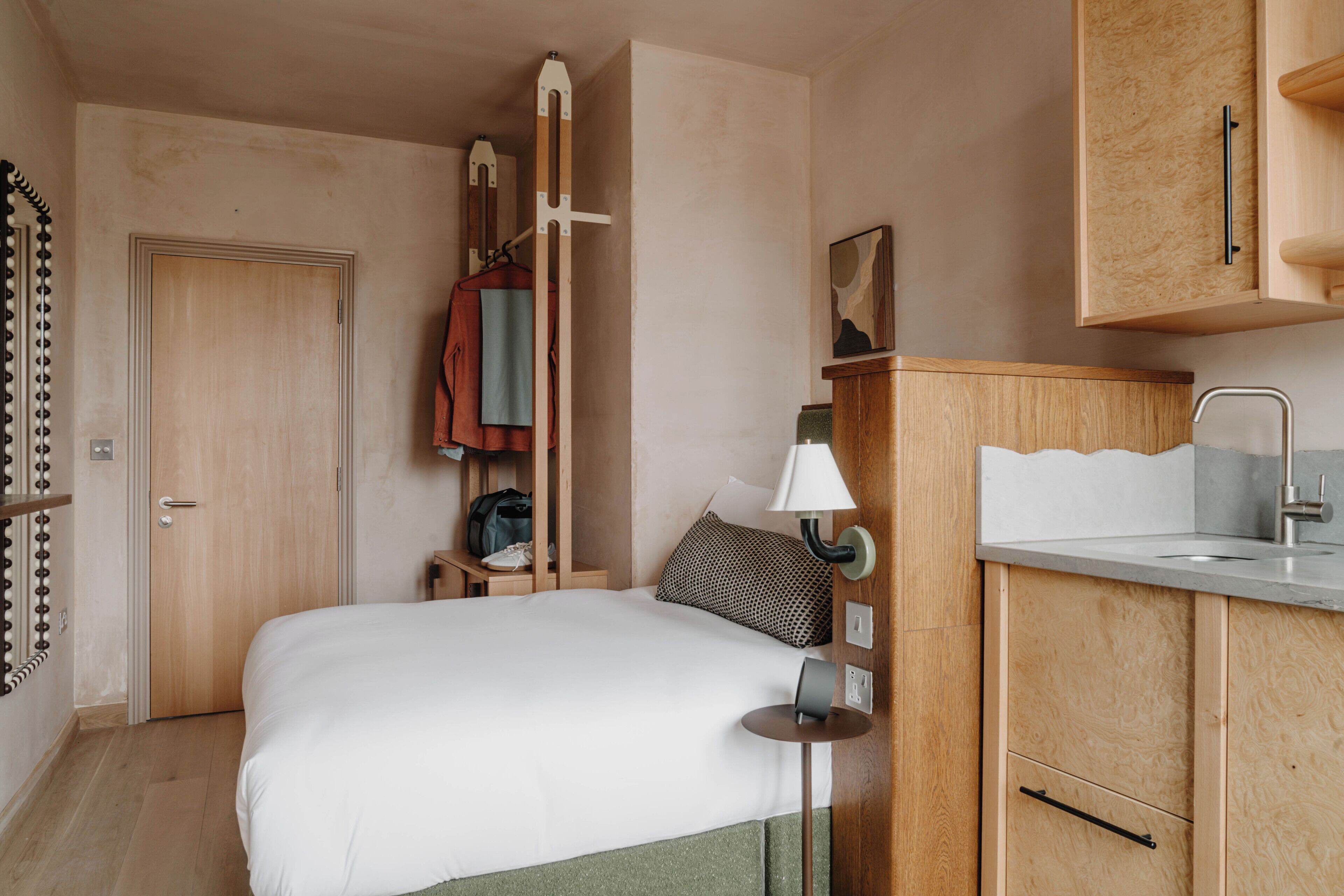 A hotel room with a bed, a wooden wardrobe, a small bedside table, a lamp, and a hanging coat rack.