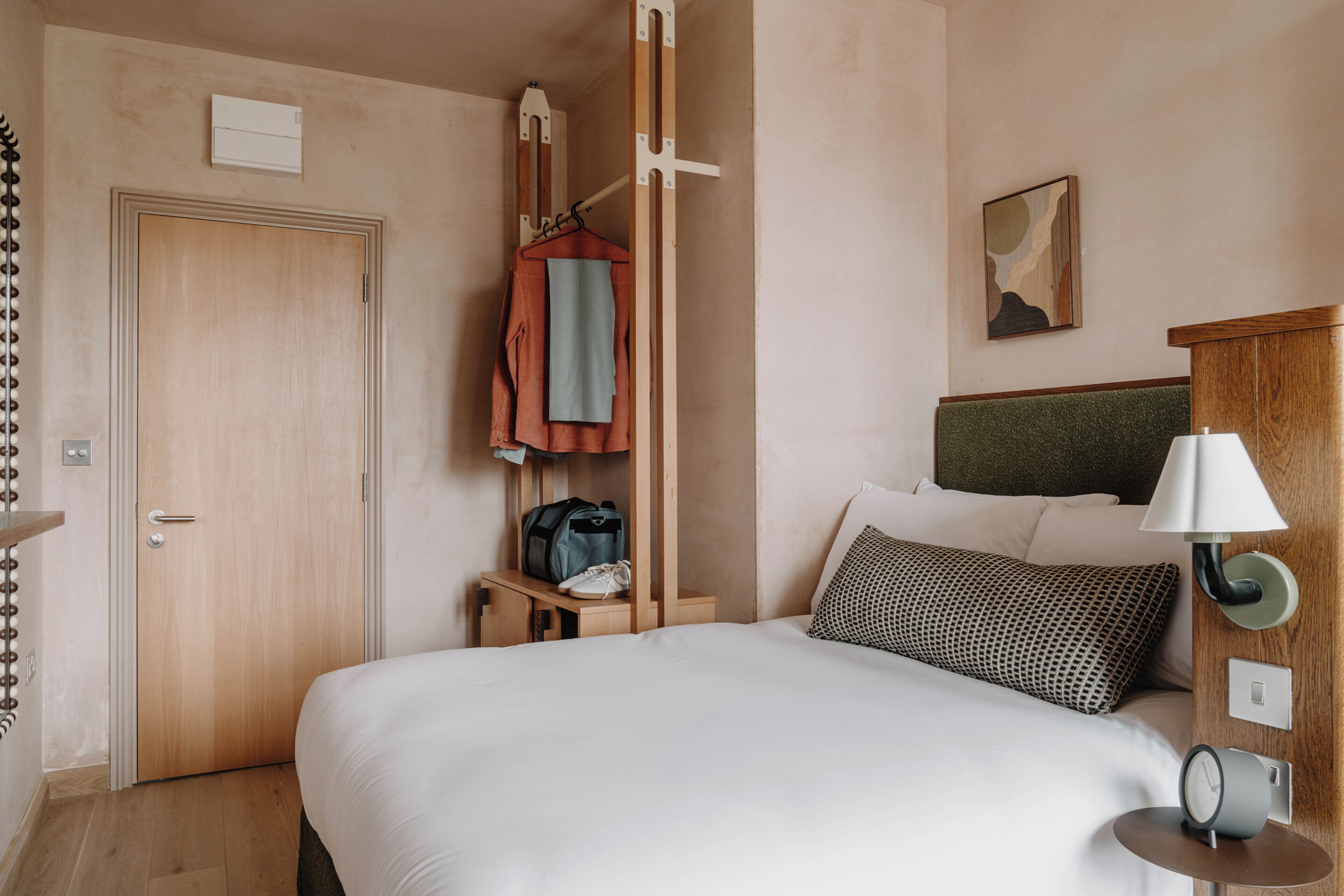 Een hotelkamer met een bed, een nachtkastje met een lamp, een hangklok, een kapstok met een jas, een deur, en een klein kunstwerk aan de muur.