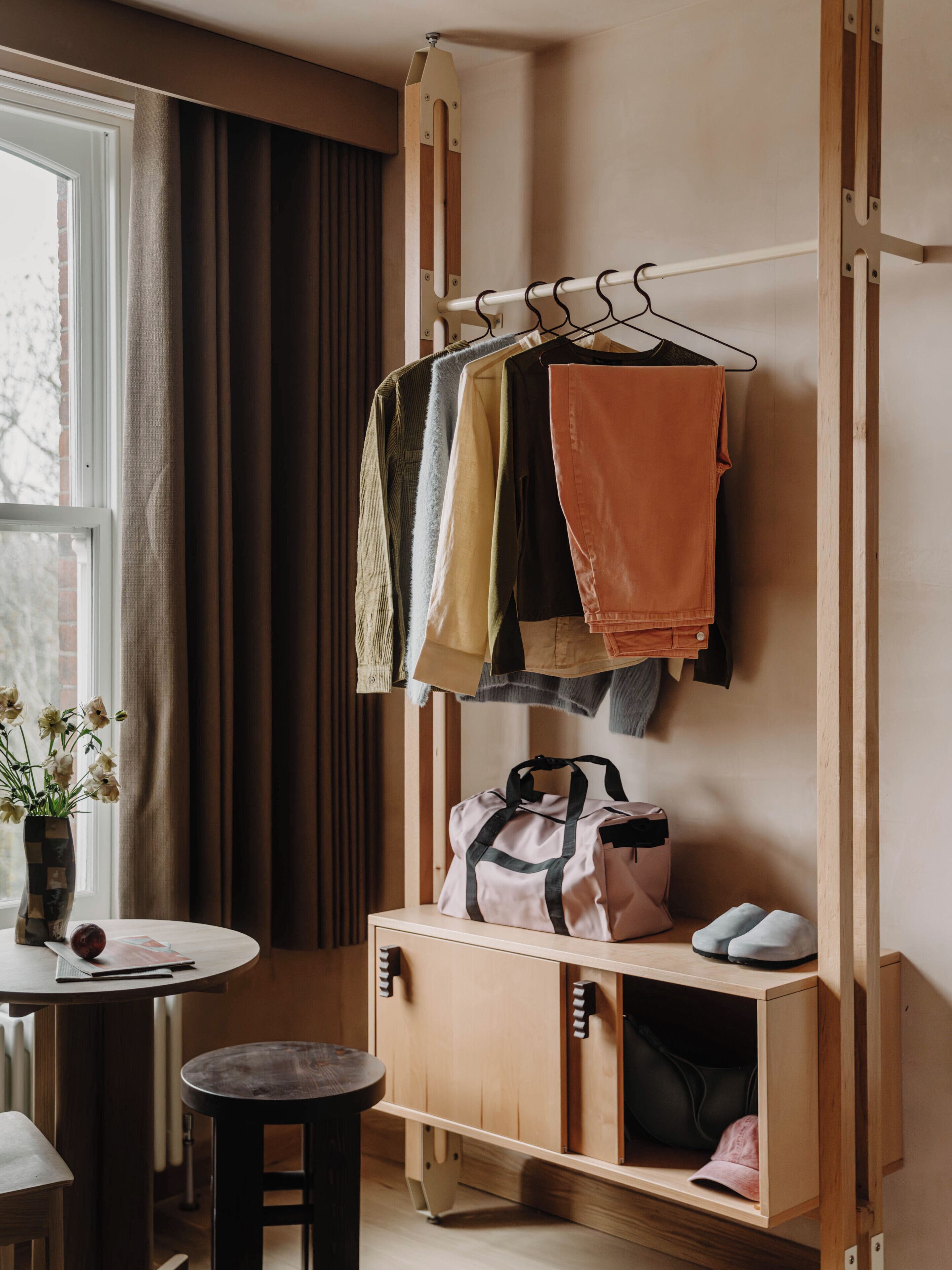 A room with a wooden wardrobe, a hanging rack with clothes, a small table with a vase, and a stool.