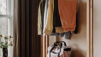 A room with a wooden wardrobe, a hanging rack with clothes, a small table with a vase, and a stool.