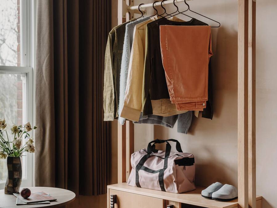 A room with a wooden wardrobe, a hanging rack with clothes, a small table with a vase, and a stool.