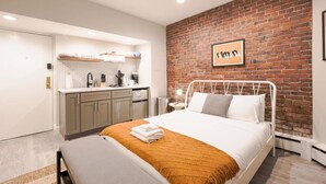 Ett modernt hotellrum med en accentvägg i tegel, en vit säng och ett köksområde med en vit bänkskiva och skåp.