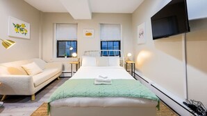 A modern bedroom with a bed, a sofa, two bedside tables, a flat-screen TV, and artwork on the walls. - Studio Nestled in Back Bay! (Boston)
