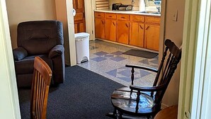 A living room with a dark carpet, a brown leather sofa, and a wooden rocking chair. A kitchen with wooden cabinets and a window is visible through an open door. - Peaceful 2-bedroom apartment with AC in charming Canaan (Canaan)