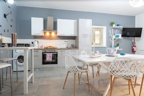 A modern kitchen with white cabinetry, a built-in oven, a washing machine, and a dining area with white chairs. - Apartment 'Oasi Giardinello' with Wi-Fi and Air Conditioning (Agrigento)