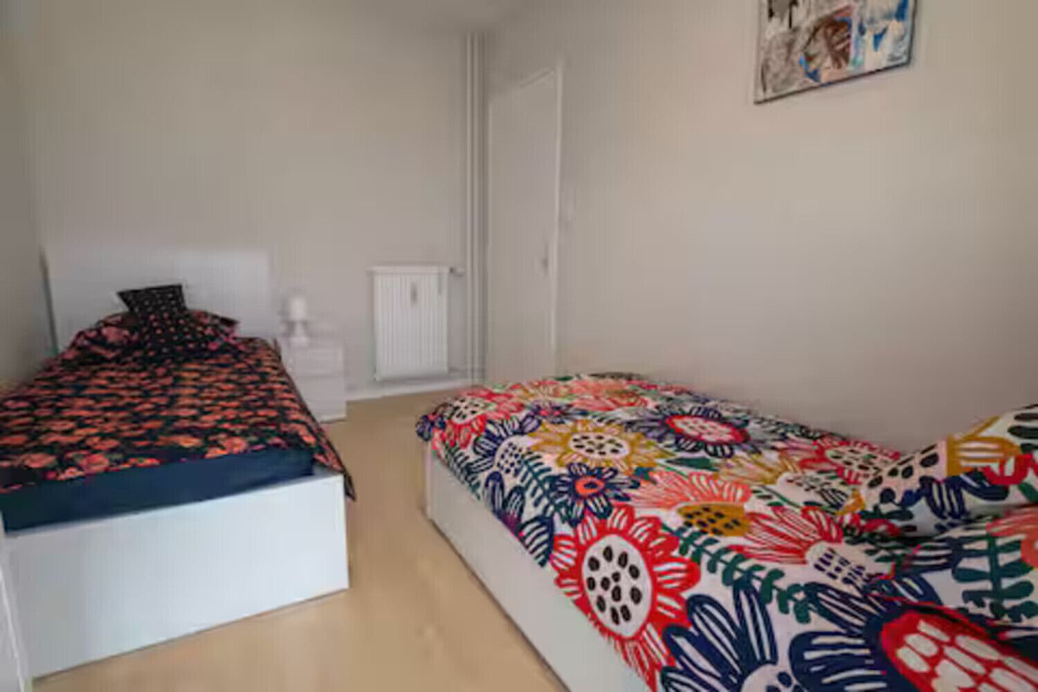 A bedroom with two beds, one with a red and black patterned bedspread and the other with a colorful floral bedspread. There is a white dresser and a radiator on the wall.