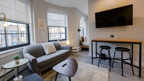 A modern living room with a sofa, a coffee table, a small side table, a TV mounted on the wall, and a dining area with stools. - Spacious Studio | Prudential Area (Boston)