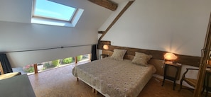 Ein Schlafzimmer mit schrägen Wänden, einem Oberlicht, einem Bett mit floralem Bettzeug, einem Nachttisch mit Lampe und Blick auf Grünfläche.