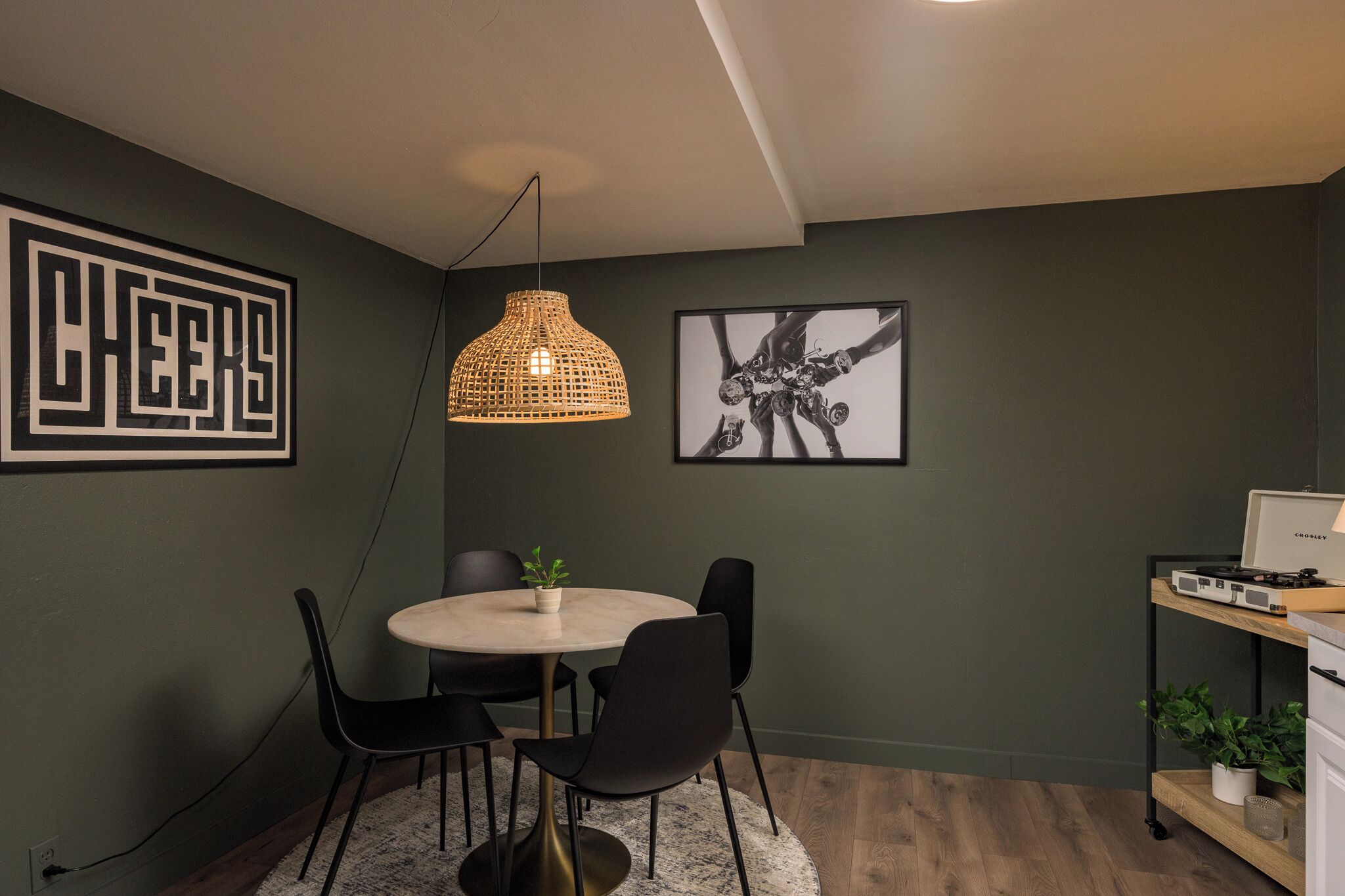Une pièce moderne avec une table ronde, des chaises noires, un luminaire suspendu en fibres tressées et une œuvre d’art abstraite encadrée.