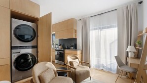 Ein modernes Hotelzimmer mit einer Küchenzeile, einer Sitzecke mit zwei Stühlen, einem großen Fenster mit Vorhängen und integrierten Geräten, darunter eine Waschmaschine und ein Trockner.