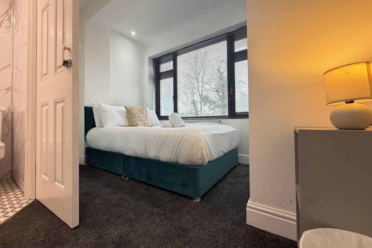 A bedroom with a bed, a window with blinds, a lamp, and a door.