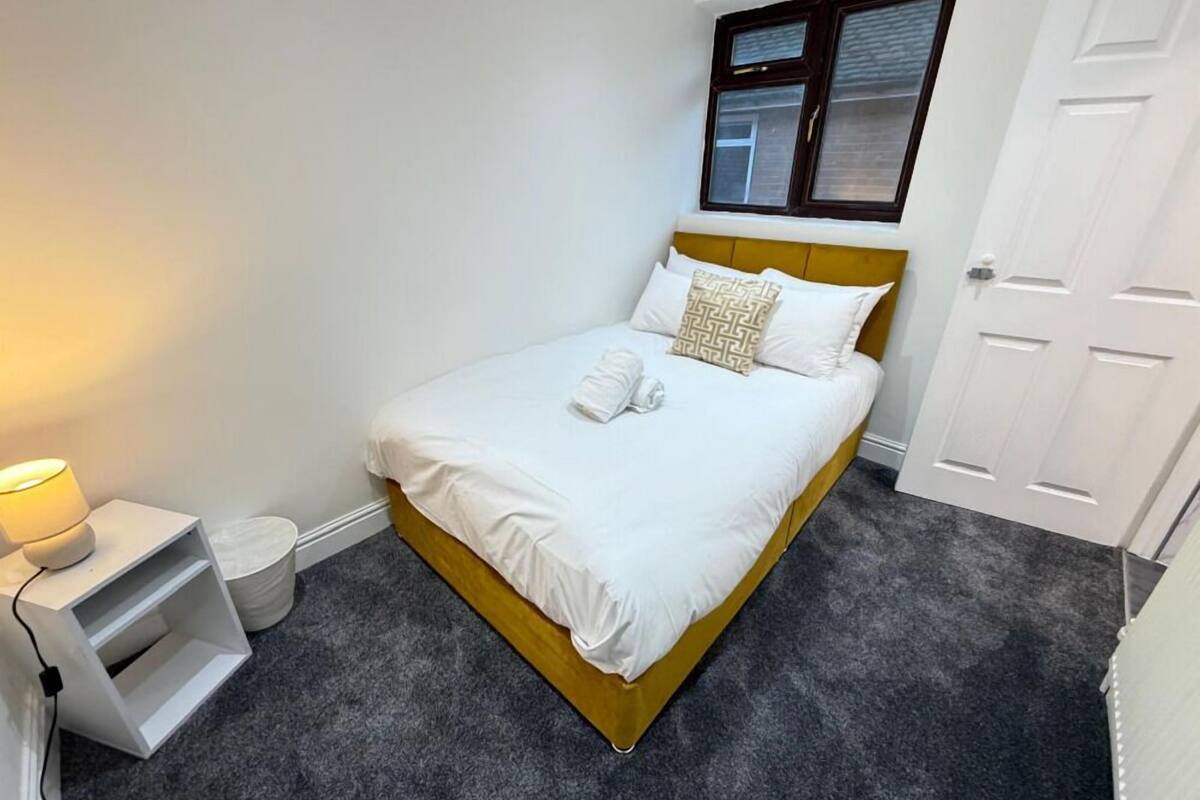 A neatly made bed with white linens and a patterned pillow, a bedside lamp, and a white nightstand with a charging cable.