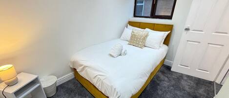A neatly made bed with white linens and a patterned pillow, a bedside lamp, and a white nightstand with a charging cable.