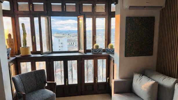 Een hoekappartement met uitzicht op de stad, een houten balkon, een stoel, een bank en een schilderij aan de muur.