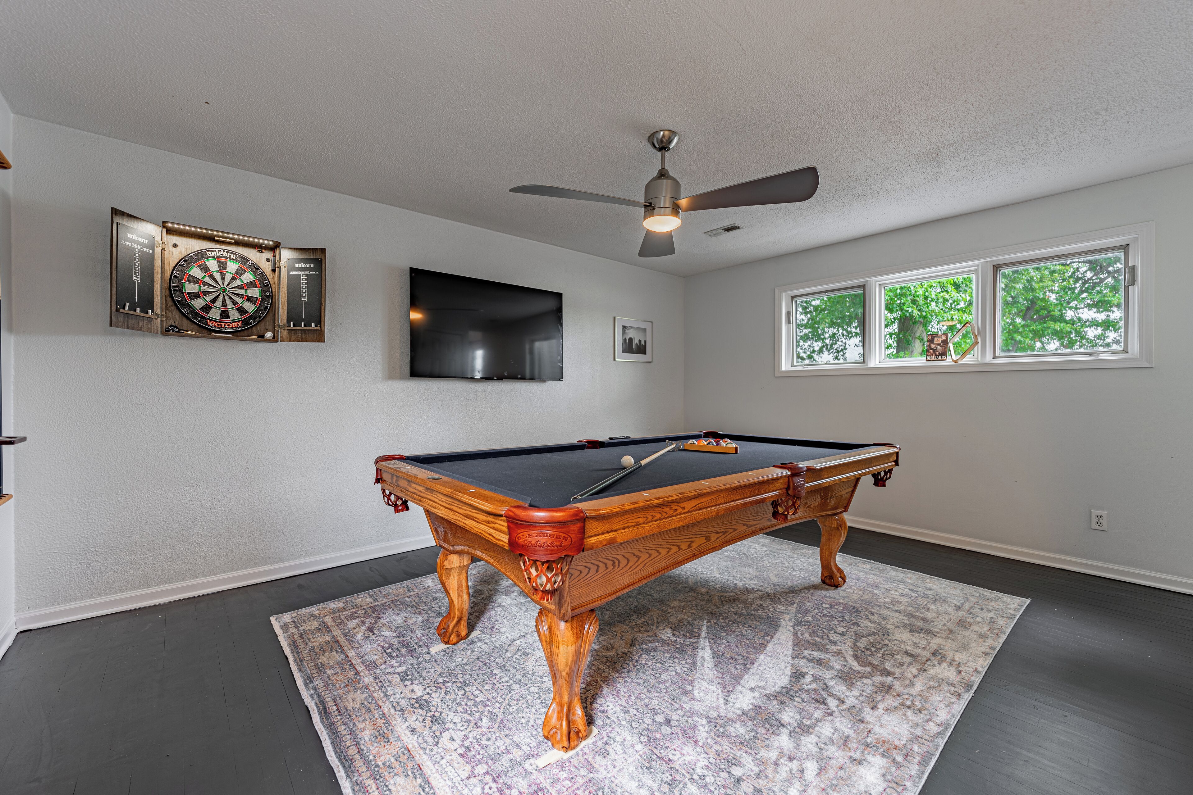 A pool table in a room with a ceiling fan, a dartboard, and a TV on the wall.