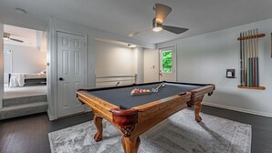 Une table de billard dans une pièce dotée d’un ventilateur de plafond, d’une porte donnant sur une autre pièce et d’une fenêtre offrant une vue sur la verdure.