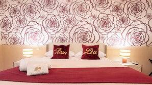 Una camera d'albergo con un letto grande, due cuscini con nomi, una coperta rossa, una parete con un motivo floreale e due lampade da comodino.