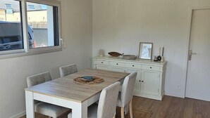 Een eethoek met een houten tafel en vier stoelen, een wit kastje met een kom en een ingelijst fotokader, en een raam met uitzicht op gebouwen.