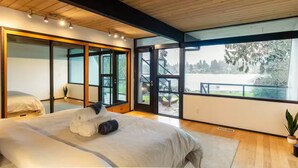 A modern bedroom with a large bed, a sliding glass door leading to a balcony, and a view of a lake. - Seattle Lake Front Oasis (SeaTac)