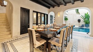 Un comedor con una mesa larga dispuesta para seis personas, rodeada de sillas, con una piscina y zona de estar exterior al fondo.