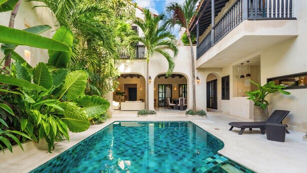 Una piscina con diseño ajedrezado, rodeada de exuberante vegetación y un balcón con barandilla.