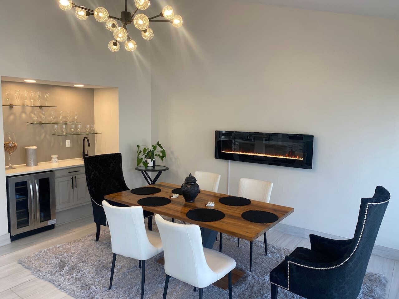 A modern dining area with a wooden table, white chairs, and a black accent chair. A kitchen with a built-in oven and a TV mounted on the wall are visible.