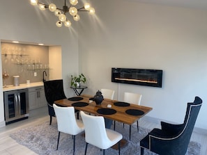 Un comedor moderno con una mesa de madera, sillas blancas y una butaca de color negro. Se ve una cocina con horno empotrado y un televisor montado en la pared.