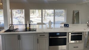 A modern kitchen with white cabinets, stainless steel appliances, a marble countertop, and large windows with blinds.