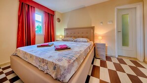 A bedroom with a bed, a nightstand, a dresser, a window with red curtains, and a door with a green panel. - Apartman ZIS 1L (Opština Bar)
