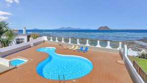 Una piscina con borde ondulado, rodeada por una solana empavedada, con vista al océano y una montaña distante.