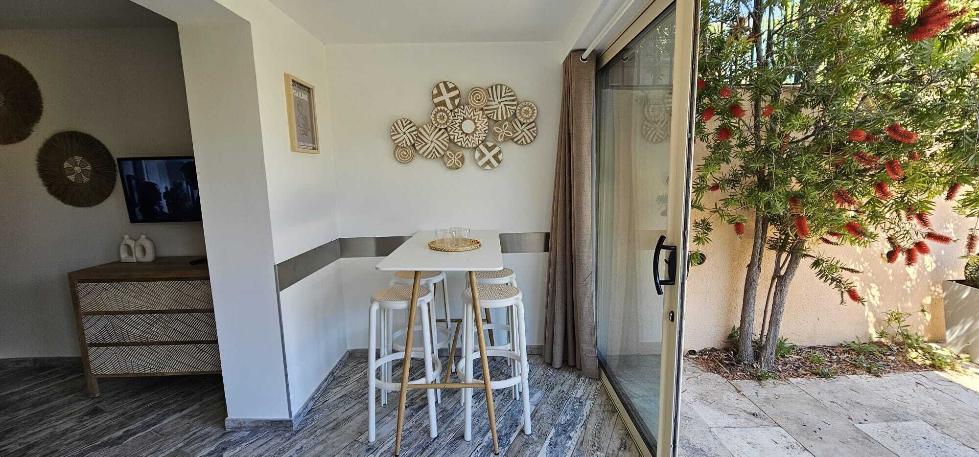 A small room with a white table and bar stools, a decorative wall, a sliding glass door leading to a garden with a tree.
