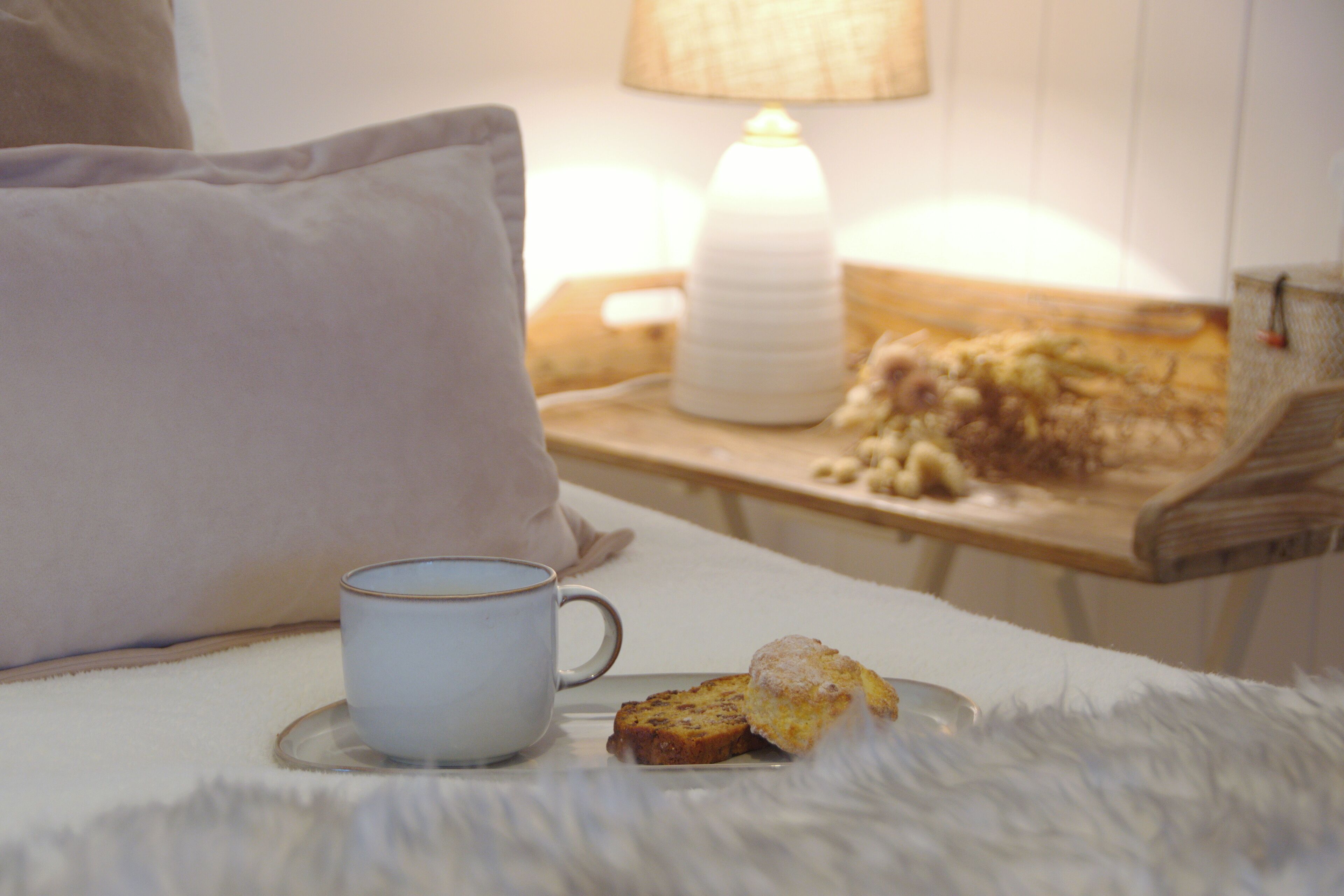 A cozy bedroom with a bed, a bedside table with a lamp, a cup, and a plate with food.