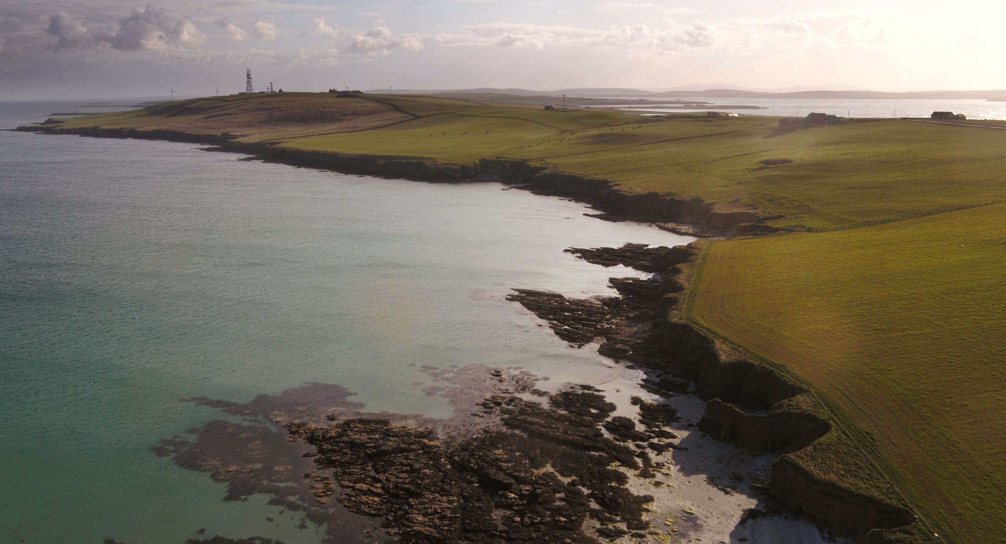 An aerial view of a coastal landscape with grassy fields, a rocky shoreline, and distant structures.