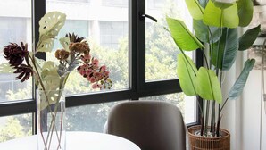 Habitació amb una finestra gran, una taula blanca, una cadira gris i dues plantes en testos.