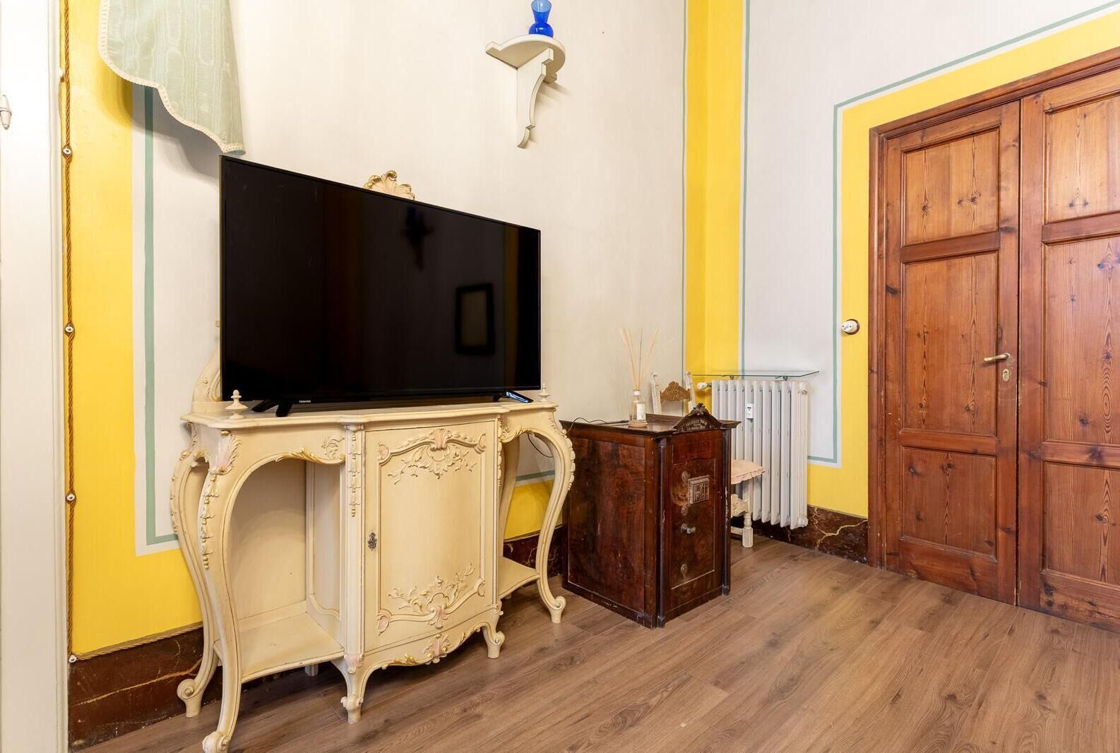 A room with a large flat-screen TV on a cream-colored cabinet, a wooden door, and a radiator.