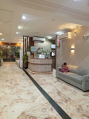 Un hall d’hôtel avec un comptoir d’accueil, des unités de climatisation, un canapé et un enfant assis dessus.