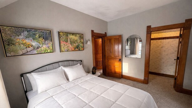 A bedroom with a large bed, wooden doors, and two framed floral paintings on the wall. - 15 Min to Lansing • 2BR • In-Unit Washer & Dryer (Grand Ledge)