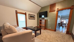 A living room with a beige sofa, a coffee table, a TV, and a bookshelf. - 15 Min to Lansing • 2BR • In-Unit Washer & Dryer (Grand Ledge)