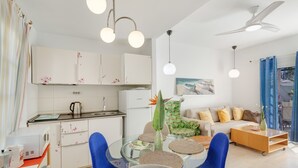 Eine moderne Küche mit weißen Schränken, einem Esstisch mit blauen Stühlen und einer Wohnbereits mit einer Couch und einem Bild an der Wand.