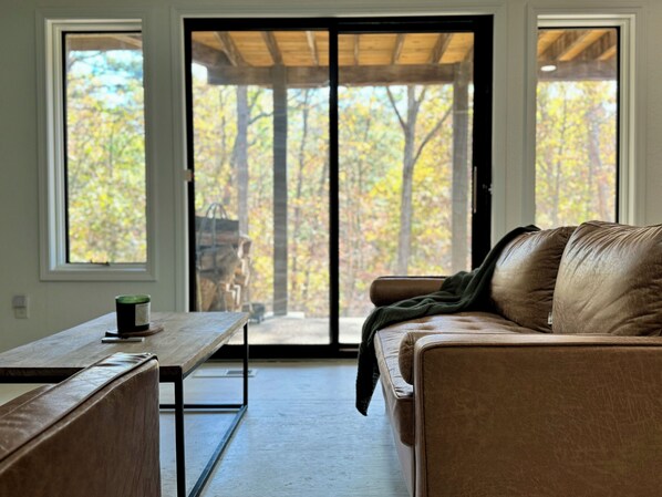A living room with a brown leather sofa, a wooden table, and a bench. There are two windows with a view of trees and a wooden structure outside.
