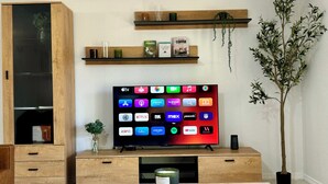 Un saló modern amb un televisor de pantalla plana sobre un suport de fusta, una taula de cafè amb dues tasses de cafè, una planta en test i una prestatgeria amb llibres i una tassa de cafè.