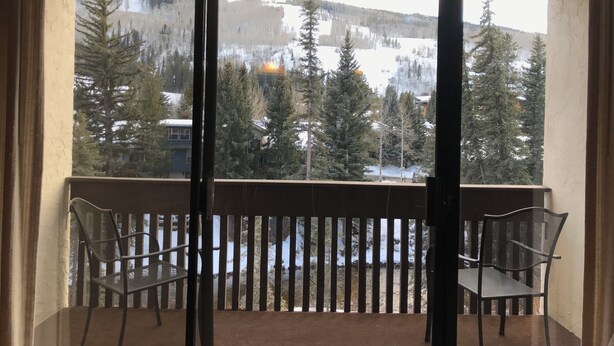 En balkon med to metalstole, udsigt til et sneklædt bjerglandskab, og en bygning i baggrunden.