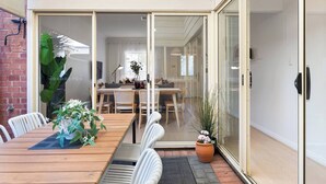 Un comedor moderno con una mesa y sillas de madera, un vidrio corredizo que da a una sala de estar, y una planta en maceta a la derecha.