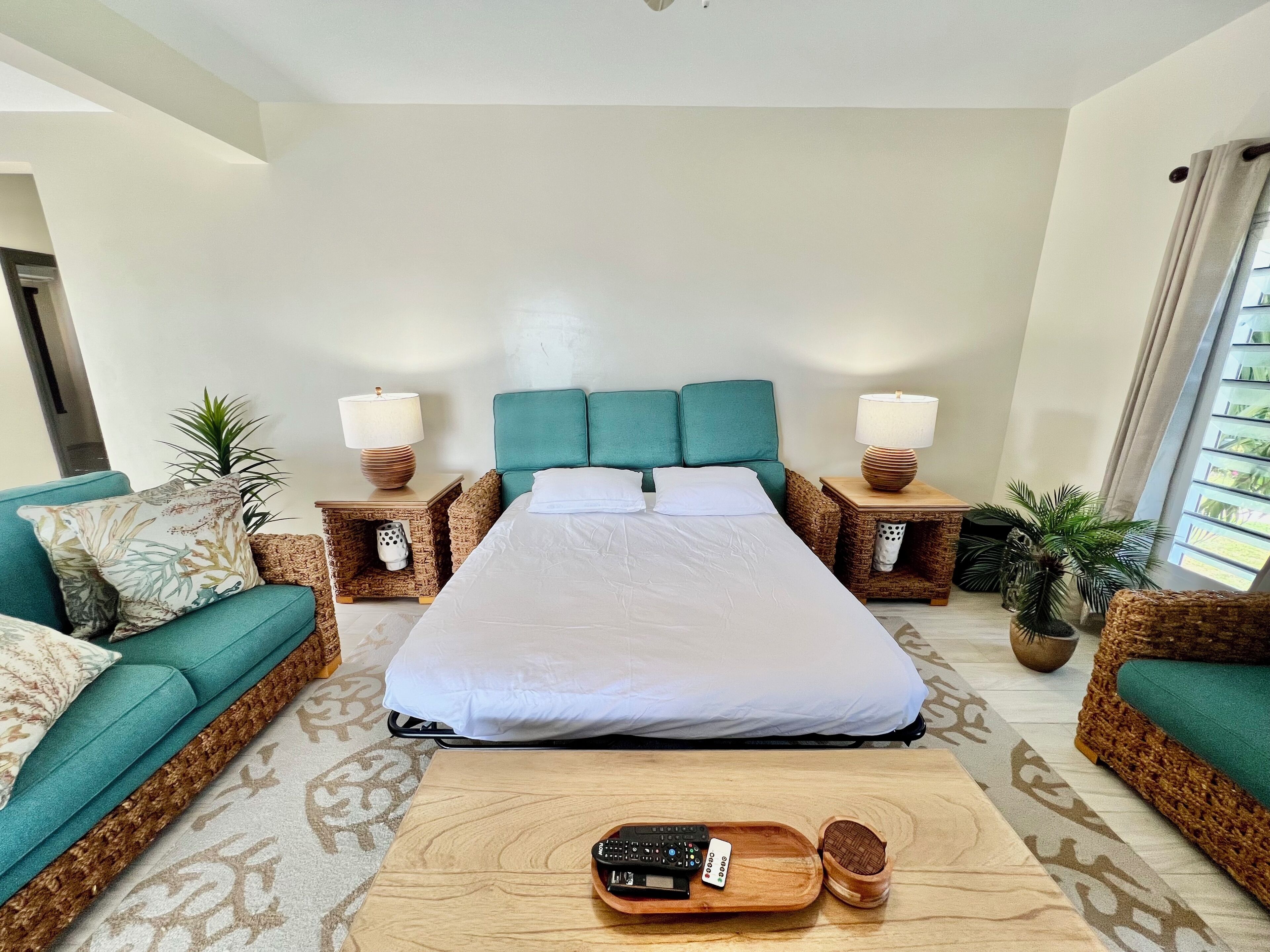 A hotel room with a large bed, two nightstands with lamps, a teal sofa, a patterned rug, and a wooden coffee table with a remote control.