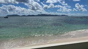 En strand med klart turkis vand, en båd i det fjerne og fjerne bjerge under en let skyet himmel.