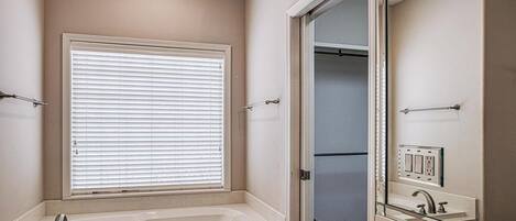 Une salle de bain avec une baignoire, une fenêtre avec des stores, un miroir et un dressing.