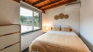 Ein Schlafzimmer mit Holzbalkendecke, einem Bett mit gemusterten Bettbezügen, einem Nachttisch mit Lampe und einem Fenster mit Blick ins Freie.