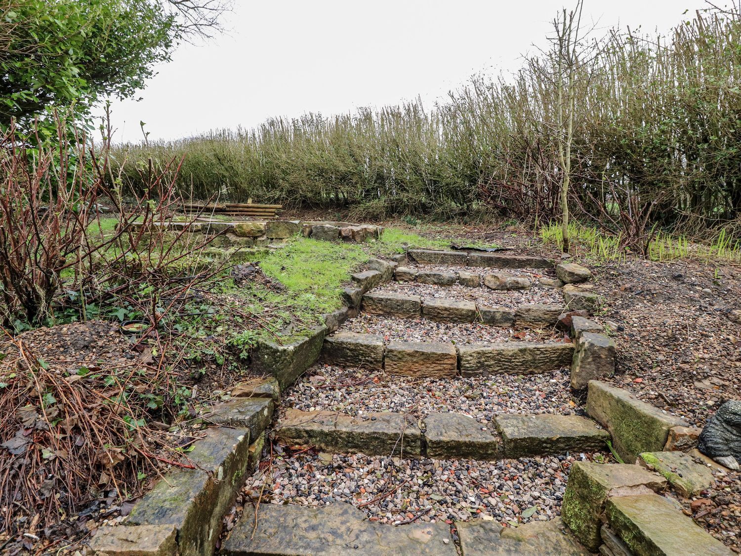 A stone pathway leading through a natural area with dense vegetation and a stone wall in the background.