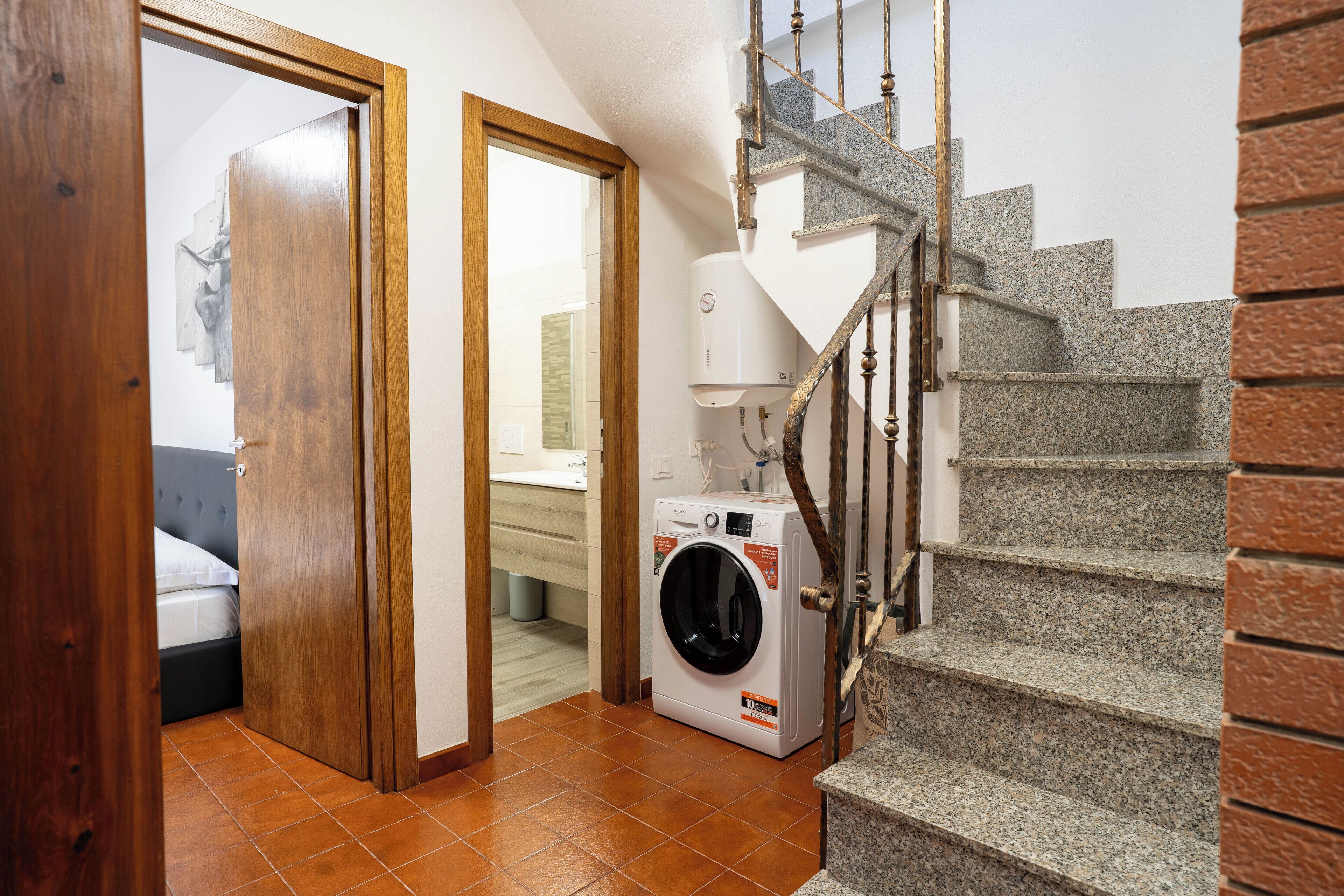 Un cuarto de lavandería con una lavadora, una puerta de madera que da a un dormitorio y una escalera con barandilla de piedra.