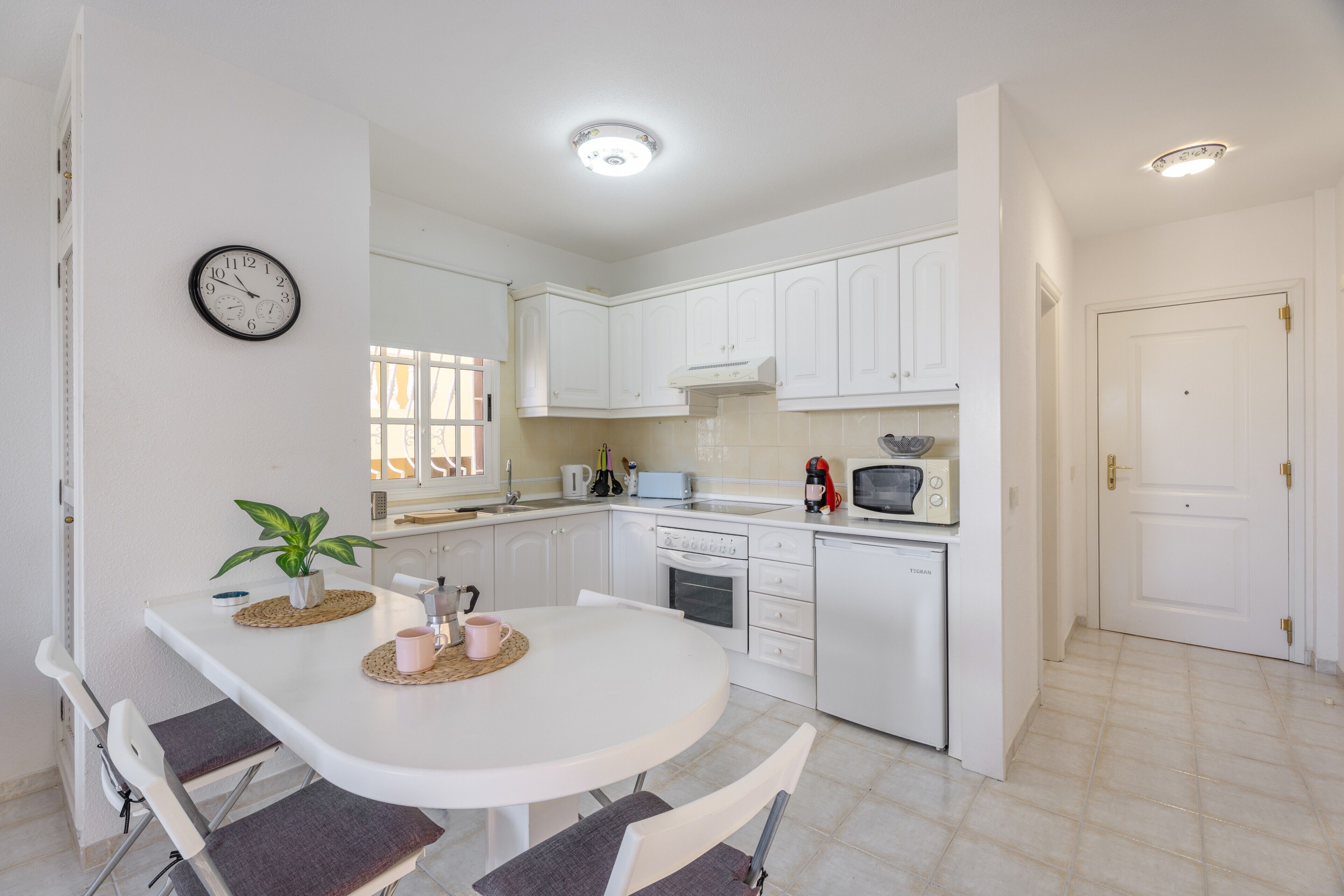 Une cuisine moderne avec des meubles blancs, une table à manger avec des chaises, une horloge et un four à micro-ondes.